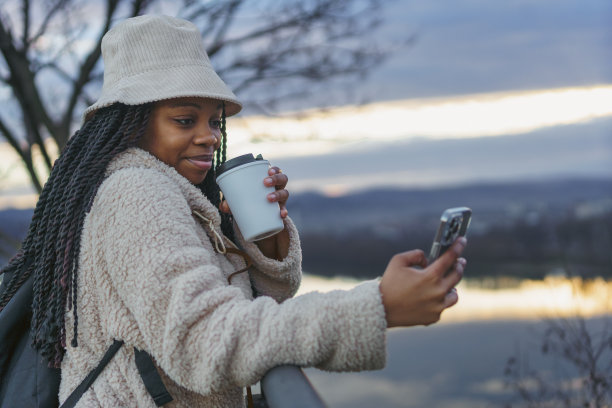 一个美丽的黑人女性对着镜头兴奋地微笑，她戴着水桶帽，背着背包，喝着咖啡，用她的手机拍照图片下载