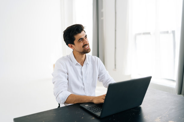 工作室拍摄的愉快的胡子年轻男子在衬衫使用笔记本电脑工作梦想看向别处坐在桌子上，室内光线。图片下载