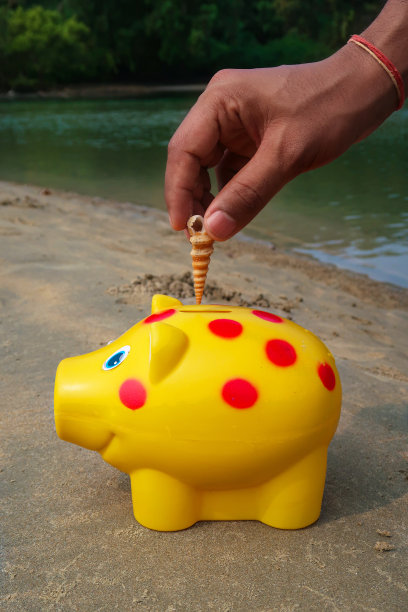 特写照片:一个认不出来的人把螺旋形的贝壳塞进塑料上的槽里，沙滩上有红色斑点的黄色储蓄罐，退潮时的海水边缘，沿海背景，假日财务和储蓄概念图片下载