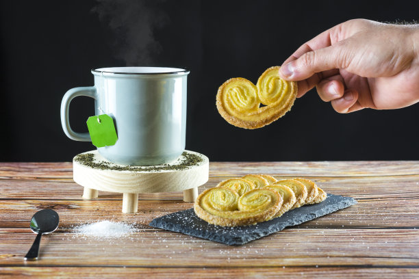 一个人的手拿着准备吃的饼干和一杯热茶。糖撒在桌上图片下载