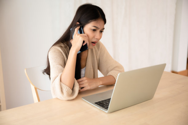 年轻的亚洲妇女一边在家里用笔记本电脑工作，一边用手机说话。在家办公，视频会议，视频通话图片下载