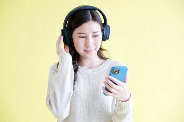 年轻的亚洲女子戴着耳机，拿着手机听音乐，站在孤立的黄色背景图片下载