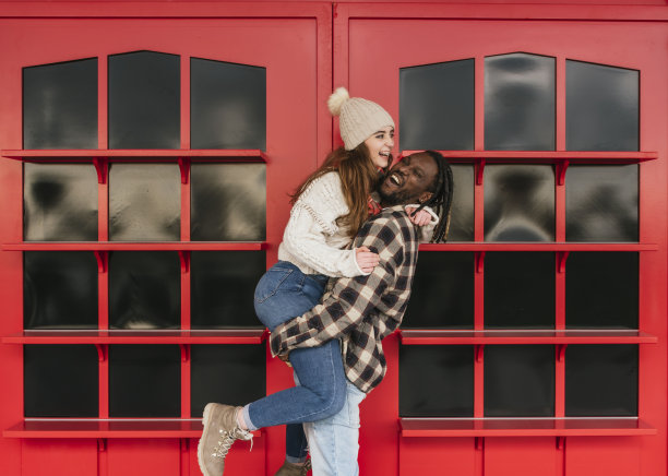 微笑的多种族夫妇在户外玩得很开心，男人抱起他的女朋友，背景是红色的门图片下载