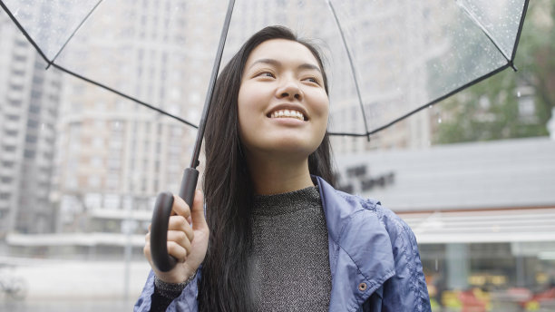 快乐的亚洲女人微笑着，享受着雨，带着透明的雨伞走在城市里图片下载