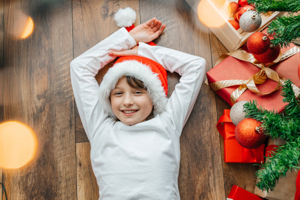 一个戴着圣诞帽的快乐女孩躺在圣诞树旁微笑着。孩子们准备庆祝新年和圣诞节。假期准备图片下载