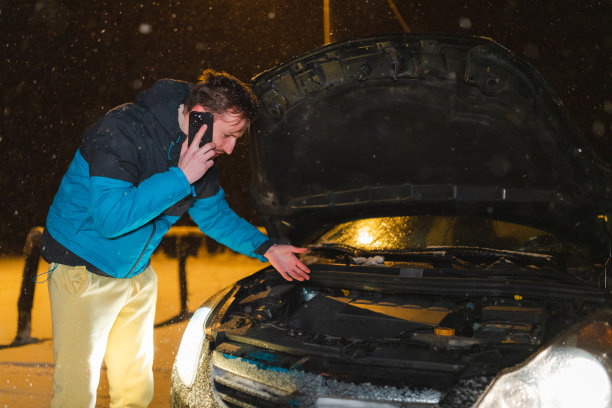 一个年轻人，被困在雪地里，一边打电话一边看他的汽车引擎盖下图片下载