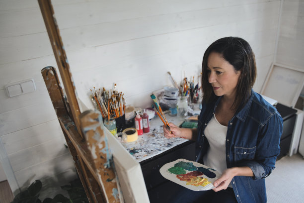 艺术、创意、爱好、工作和创意职业的概念。一个忙碌的艺术家坐在画架前的椅子上，用画笔作画，使用丙烯酸颜料。图片下载