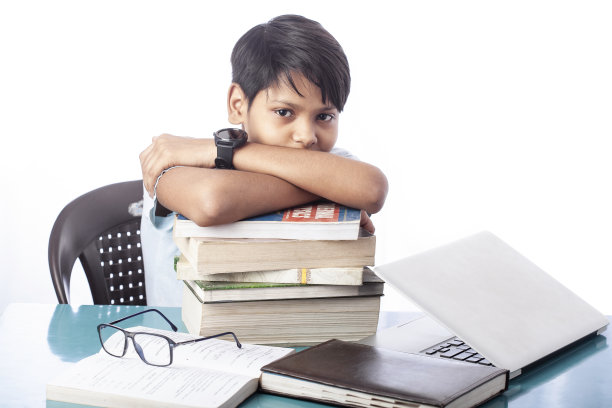 聪明的男孩看着相机躺在一堆书和笔记本电脑在桌子上图片下载