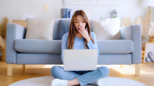 年轻的亚洲女性在家里的笔记本电脑上工作，而坐在地板上靠近沙发。兴奋的女优胜者一边看笔记本一边庆祝成功图片下载