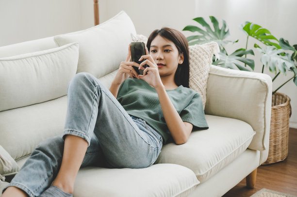 快乐的年轻亚洲女子在家里放松。女人躺在沙发上用手机图片下载