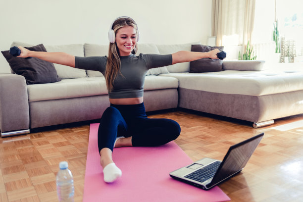 一个运动的年轻女子在家里锻炼时使用笔记本电脑和举着哑铃。图片下载