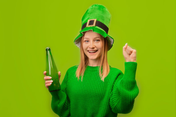 快乐的微笑女孩在圣帕特里克节妖精绿色帽子拿着绿色啤酒瓶，高兴，说，是，庆祝胜利和保持拳头泵手势隔离在彩色绿色背景。图片下载