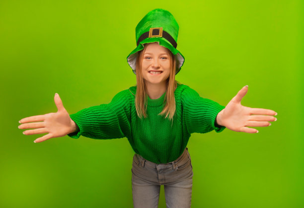 圣帕特里克节庆祝理念:身穿绿色毛衣、头戴绿帽子的小妖精女孩伸出双手自由拥抱，在彩色绿色背景上邀请拥抱、支持和欢迎孤立的人。图片下载