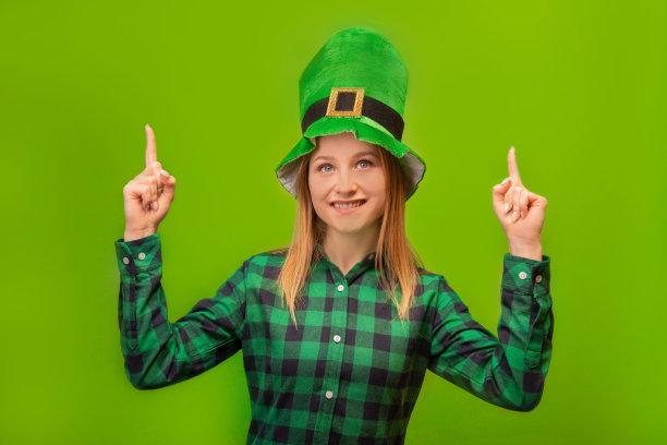 圣帕特里克节的小妖精女孩伸出食指或举起手指，穿着绿色格子衬衫和绿色传统爱尔兰帽子，孤立地看着彩色绿色背景。图片下载