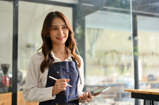 迷人的亚洲女服务员或咖啡店员工在围裙拿着平板电脑，站在她的咖啡馆。图片下载