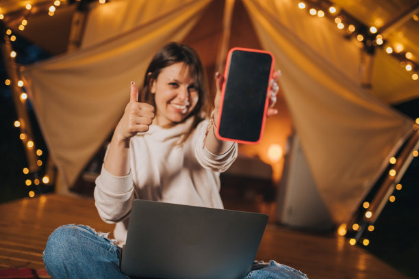 快乐的女人自由职业者在夏夜舒适的露营帐篷上工作时展示手机。户外度假、度假的豪华露营帐篷。生活方式的概念图片下载
