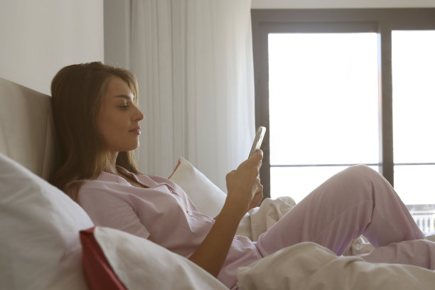 穿着睡衣，床上放着手机的年轻女人。图片下载