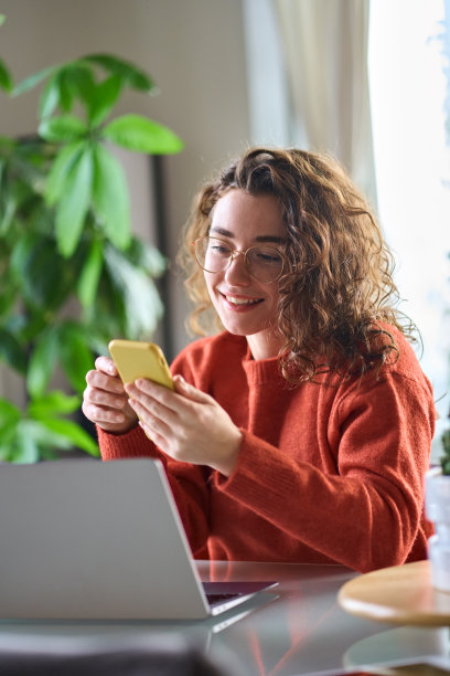 年轻快乐的漂亮女孩坐在桌子上拿着智能手机使用手机。图片下载