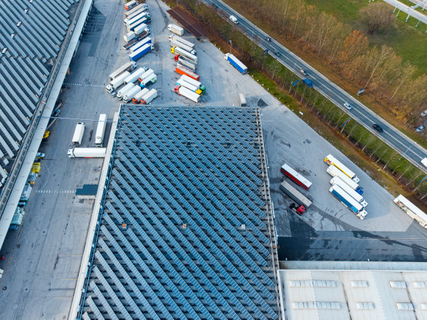 大型太阳能电池板阵列农场在一个配送仓库建筑，卡车停在外面自上而下的观点图片下载