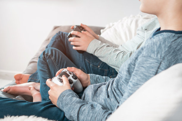 裁剪的照片，两个十几岁的男孩坐着，拿着游戏控制器操纵杆，玩游戏机站的视频游戏。图片下载