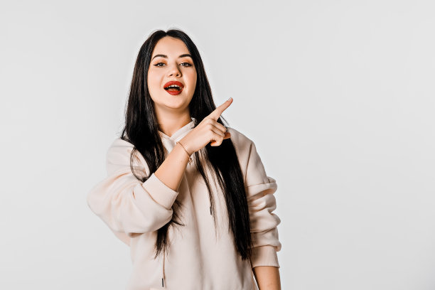 看看这个。微笑的年轻女人。美丽的黑发女孩的肖像与高兴的面部表情指着手指离开复制空间，白色的工作室背景图片下载