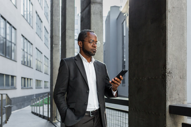 严肃而成功的非裔美国商人老板在办公楼外使用智能手机，西装革履的男子在网上阅读新闻，思考和浏览网页图片下载