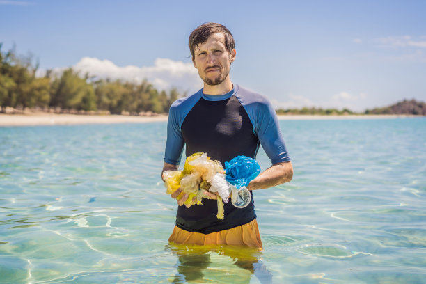 一名男子从美丽的蓝绿色大海中收集包裹。天堂海滩污染。沙滩上的垃圾垃圾洒在沙滩上造成的人为污染和环境问题，倡导志愿者概念清洁图片下载