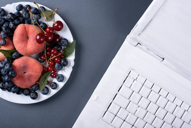 灰色背景的夏日水果和白色涂料的笔记本电脑。桃子和樱桃图片下载