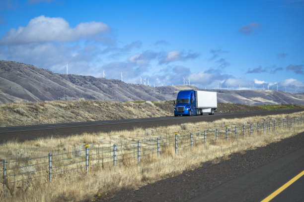 亮蓝色的大卡车在直道上用半挂车运输货物，背景是风力涡轮机图片下载