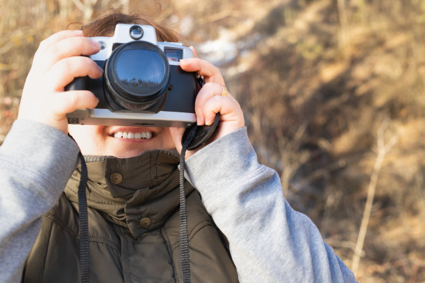 微笑的女摄影师拿着相机靠近她的脸，双手穿着温暖的照片，一个美丽的大自然在阳光明媚的冬日。图片下载