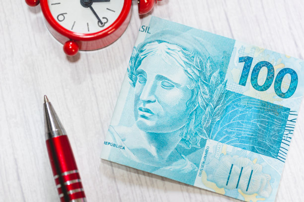 一张近距离照片中，一张价值100雷亚尔的巴西雷亚尔钞票放在一张木桌上。作文中有一支钢笔和一个闹钟。经济、投资和通货膨胀。图片下载