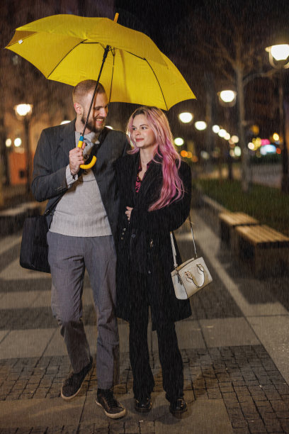 一个雨夜，一个年轻人和他的女朋友打着黄色的雨伞走在路上。图片下载