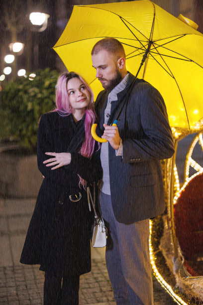 一对年轻夫妇在雨夜打着黄伞走在街上。图片下载