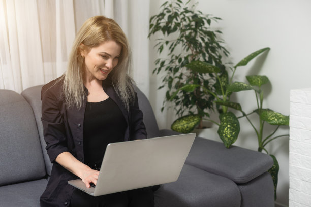 微笑的年轻女子使用笔记本电脑，坐在家里的沙发上，漂亮的女孩购物或在社交网络上聊天，玩得开心，看电影，自由职业者在电脑项目上工作。图片下载