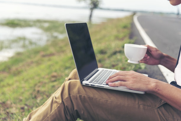 一名男子在办公室外拿着咖啡杯，手里拿着打字笔记本电脑。近距离的男子双手使用电脑笔记本电脑坐在绿色公园户外生活方式。男性使用笔记本电脑家庭办公技术图片下载
