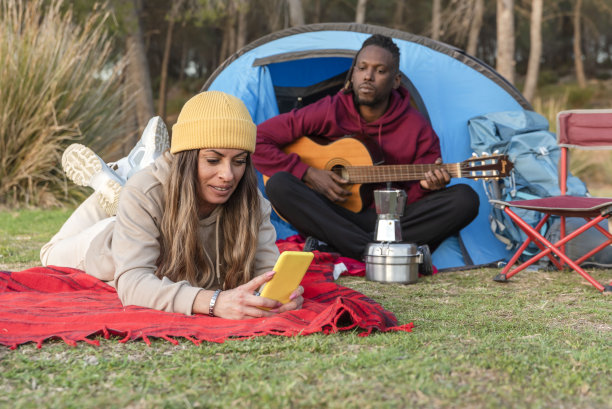 一对少数民族夫妇在露营帐篷里，女人躺在毯子上打电话，他在她身后弹吉他图片下载