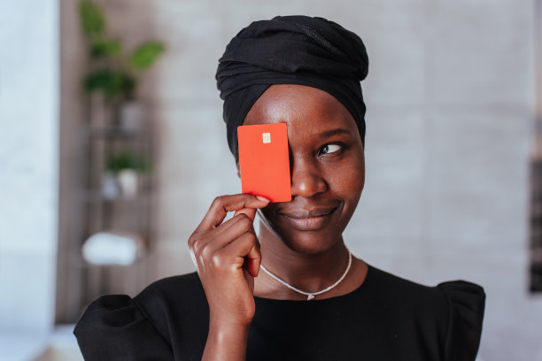 特写非洲快乐的女人拿着信用卡，遮住她的脸，笑得很宽。戴着黑色头巾和裙子的非裔美国年轻女性思考如何花钱，银行和金融。图片下载