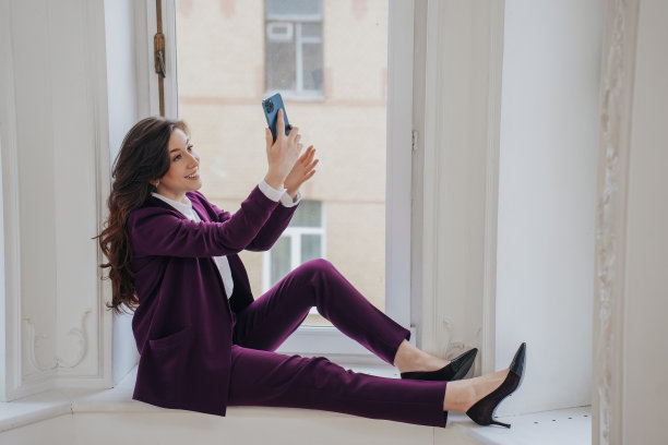 成功的棕发商人在紫罗兰西装坐在窗台上拿着电话，通过视频通话。华丽的微笑律师高兴地完成了案件。快乐的老师正在休息。图片下载