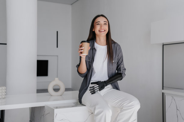 迷人的黑发白人女孩在灰色衬衫，白裤子喝咖啡，学习如何使用人工仿生网络手坐在家里。创伤后康复。年轻女子探索网络肢体。图片下载