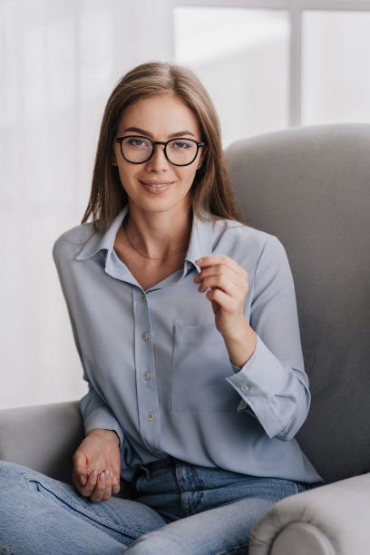 垂直拍摄自信的意大利金发年轻女子坐在椅子上，戴着眼镜，穿着蓝色衬衫和蓝色牛仔裤，微笑着看着家里的相机。迷人的瑞典家庭主妇在周末放松。图片下载
