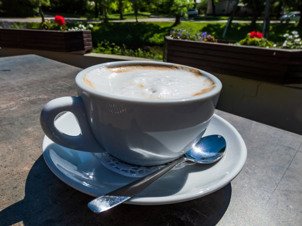 夏日，在明亮的阳光下，户外露台上的黑色桌子上放着一杯白色的卡布奇诺咖啡图片下载