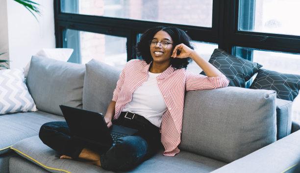 快乐的黑皮肤女人在光学眼镜对着相机微笑，而在舒适的客厅休息，快乐的非裔美国女性的肖像与现代笔记本电脑工作坐在家里的沙发上图片下载