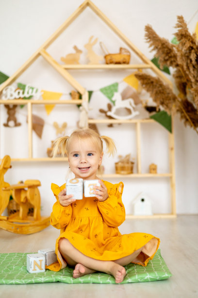 一个小女孩微笑着，摇摇晃晃地站在玩具房里的金字塔或木块塔上，她是一个穿着黄色连衣裙的快乐的孩子图片下载