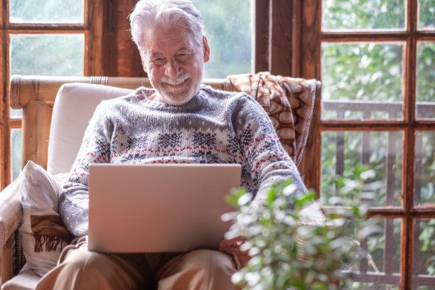 微笑放松的退休男子坐在家里，同时使用笔记本电脑上网，网上购物，看视频。年长的微笑的白人男子在山间小屋图片下载
