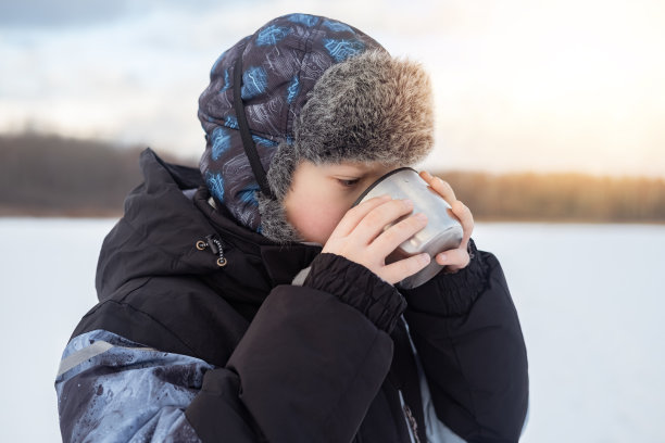 在阳光明媚的冬天，穿着暖和衣服的男孩在户外喝着热水瓶里的热茶。图片下载
