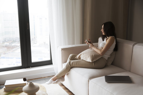 年轻女子用手机坐在沙发沙发客厅感到放松舒适。技术与人的概念图片下载