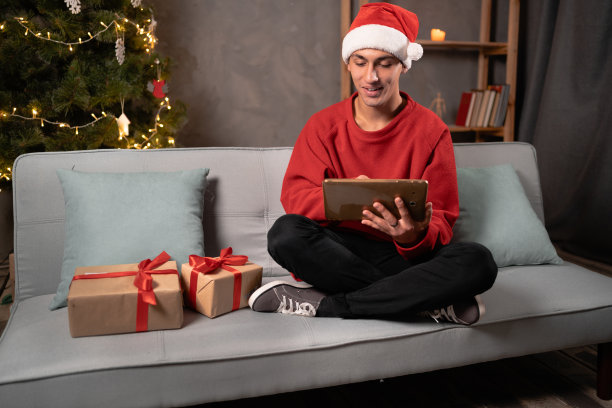 快乐的人在圣诞老人的帽子坐在沙发上与平板电脑在家里在晚上圣诞树灯的背景。人、科技和假期图片下载