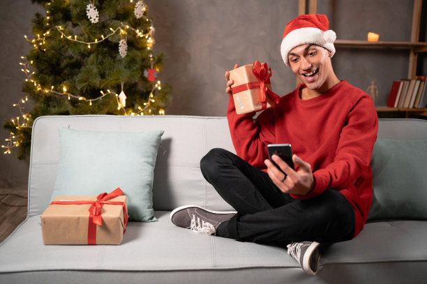 年轻快乐的男博主戴着圣诞帽，在智能手机摄像头上展示圣诞前夜坐在家里的沙发上。积极的人对圣诞礼物感到高兴图片下载