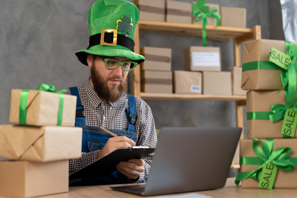 快乐的小精灵男子使用笔记本电脑和剪贴板坐在车间桌子上圣诞日。电子商务网站假日网上购物大甩卖图片下载