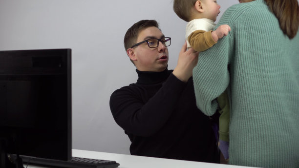 一位年轻的IT专家带着小儿子在电脑前工作。一个女人把她的儿子交给一个在家里远程工作的丈夫。一个戴眼镜，穿黑色高领毛衣的男人。图片下载